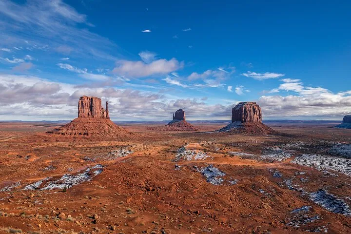 Three red rock buttes in Monument Valley under a partly cloudy blue sky.