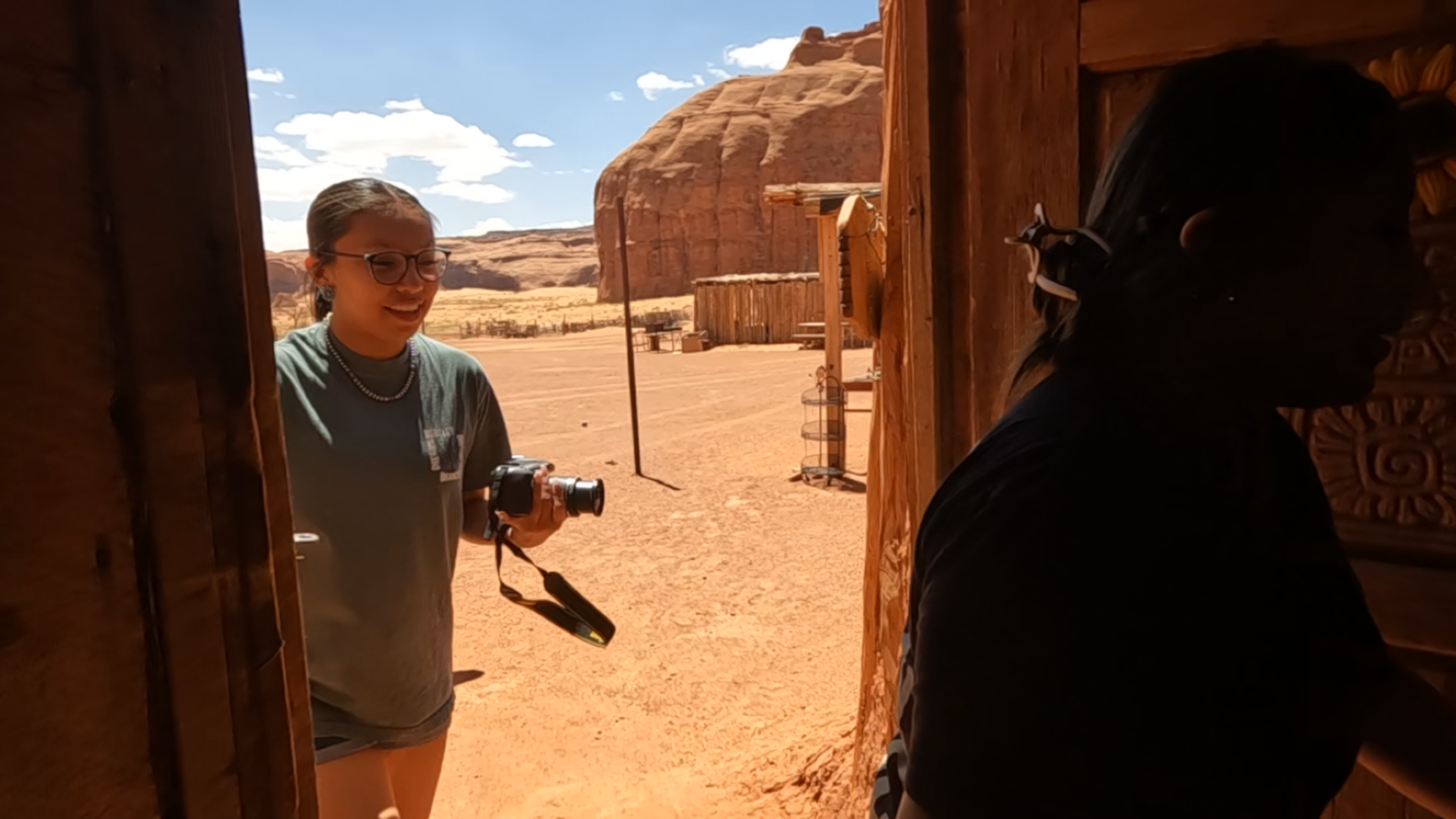 Two people, one holding a camera, at a wooden doorway in a desert area with red rock formations.