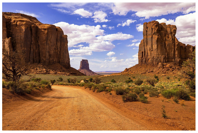 Dirt road between large rock formations under a blue sky with clouds.
