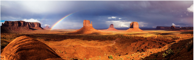 Panoramic view of desert landscape with mesas and a rainbow in the sky.