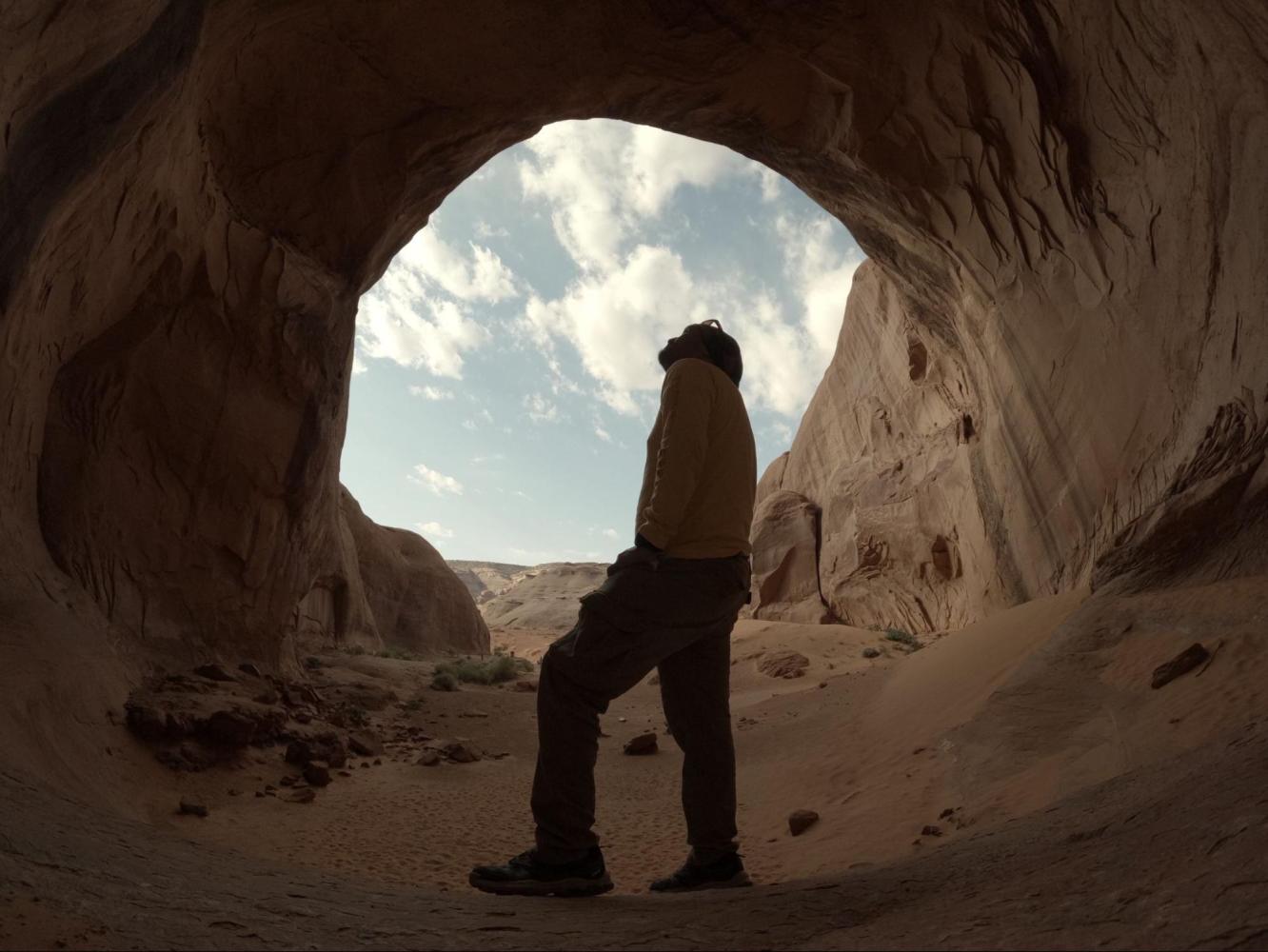 Silhouette of a person standing in a cave entrance against a cloudy sky.